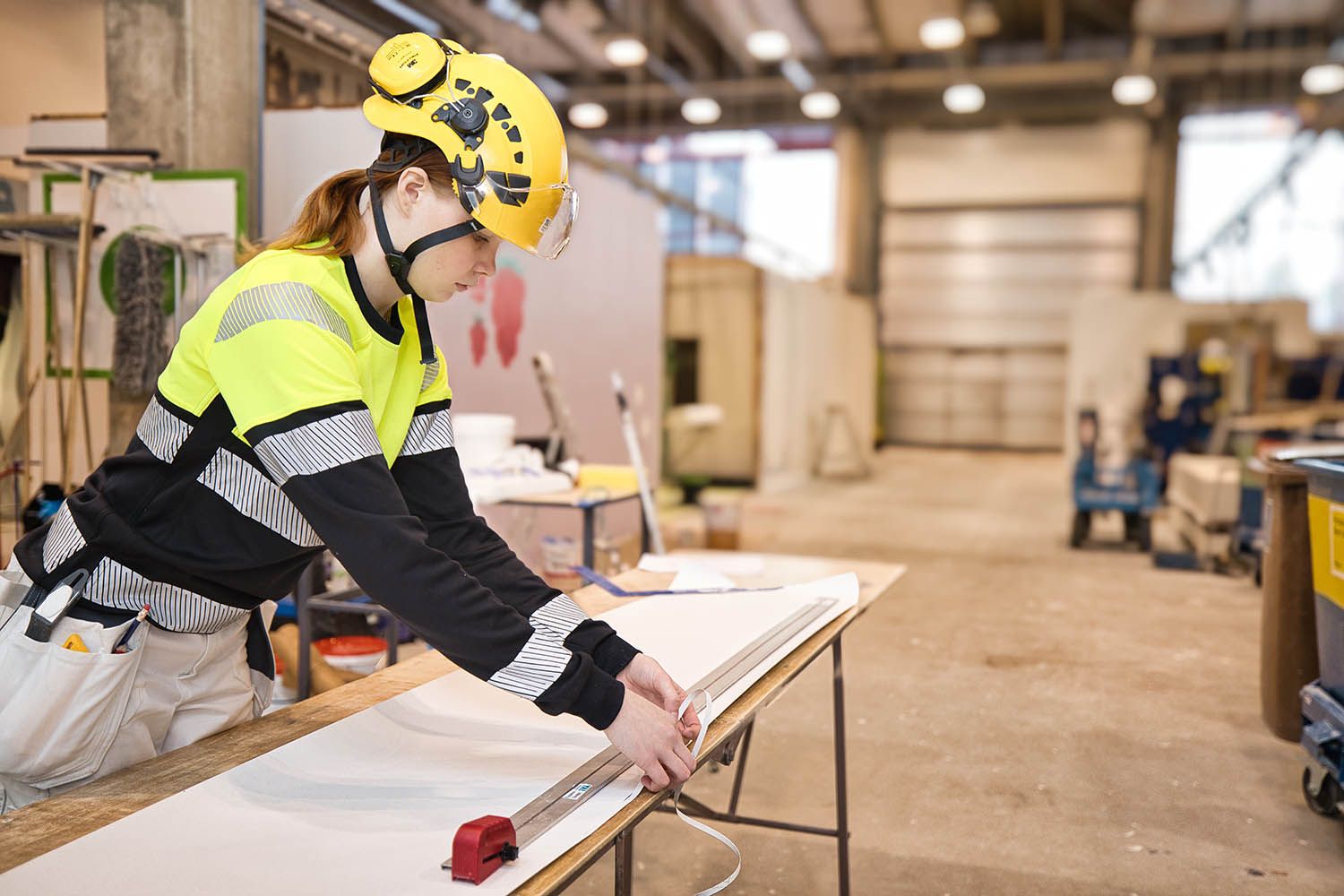 En ung ytbehandlare skär tapeter på ett bord i en bygghall